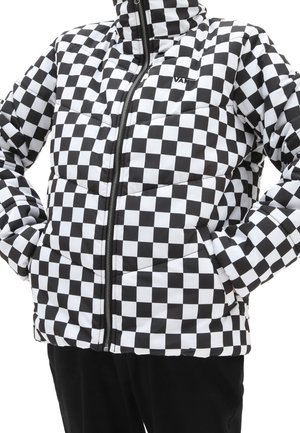 Doudoune à carreaux noir et blanc avec fermeture éclair frontale et mains dans les poches, portée sur un pantalon noir.