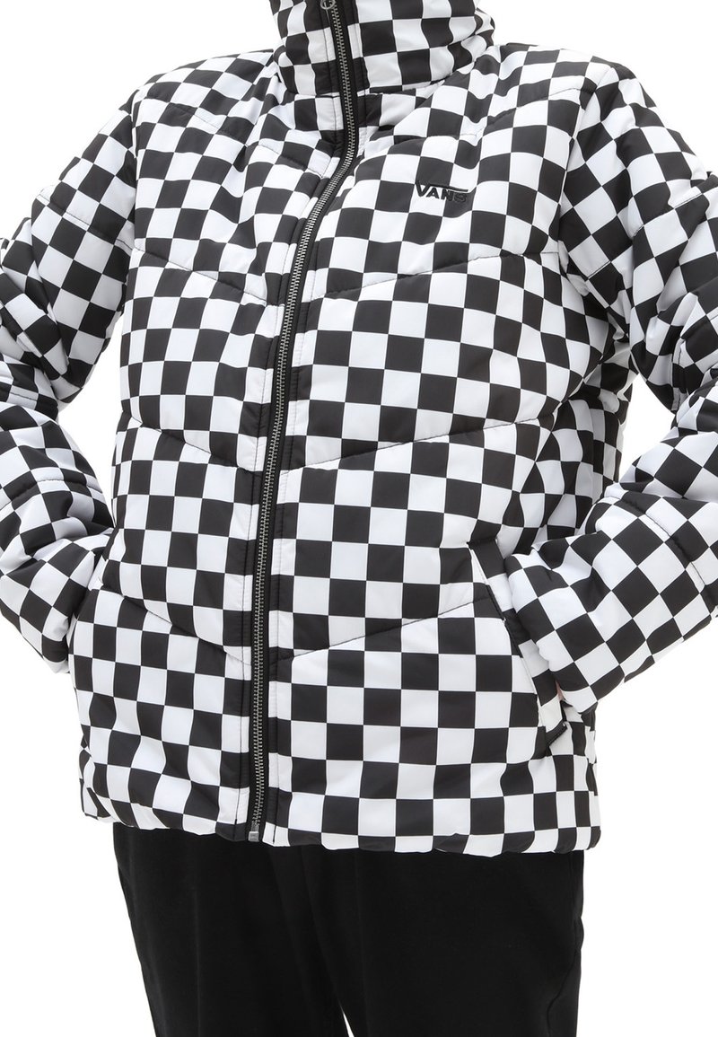 Doudoune à carreaux noir et blanc avec fermeture éclair frontale et mains dans les poches, portée sur un pantalon noir.