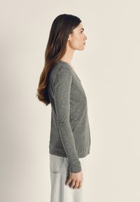 Moteris su ilgais rudos spalvos plaukais, vilkinti pilką ilgomis rankovėmis marškinėlį ir šviesius džinsus, stovinti šonu priešais paprastą šviesią fono sieną.