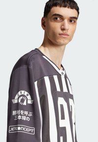 Mustvalge triibuline spordijerseys halli taustaga, millel on logod ja tekst varrukatel. Valmistatud hingavast materjalist, ümar kaelus.