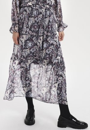 Jupe longue, transparente et à motifs présentant des designs floraux dans des nuances de violet, blanc et noir. Associée à des chaussures noires et des collants.