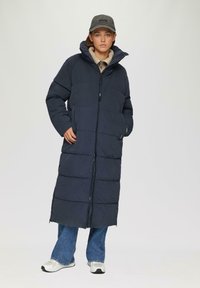 Marineblauer Puffermantel mit hohem Kragen, gestepptem Design, Reißverschluss vorne, zwei seitlichen Taschen und langer Silhouette. Kombiniert mit hellen Jeans und Sneakers.