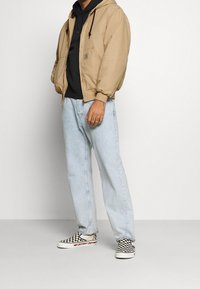 Brązowa kurtka z kapturem na czarnej bluzie, w połączeniu z jasnoniebieskimi dżinsami o prostym kroju i czarno-białymi slip-onami w kratkę.