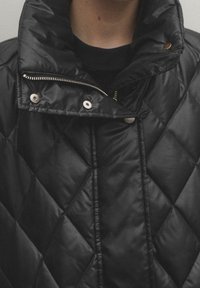 Zbliżenie osoby noszącej czarną pikowaną kurtkę z podniesionym kołnierzem, widocznym zamkiem błyskawicznym i zatrzaskami.