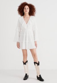 Bela ruffled mini obleka z dolgimi rokavi, v-izrezom in plastovitim dizajnom. Združena z črnimi in kremnimi kavbojskimi škornji do kolen.