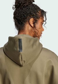 Olivengroene hoodie met een gestructureerde afwerking, voorzien van een trekkoordkap en een zwart label aan de achterkant. Krullend haar zichtbaar aan de bovenkant.