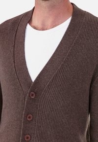 Cardigan marron côtelé avec un col en V, doté de quatre boutons foncés, porté sur une chemise blanche. Texture douce et coupe ajustée.