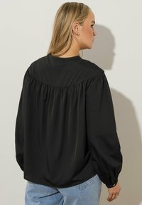 Svart långärmad blus med en rynkad baksidedesign, tillverkad av slät tyg. Har rund halsringning och knäppta ärmslut. Matchas med ljusa jeans.