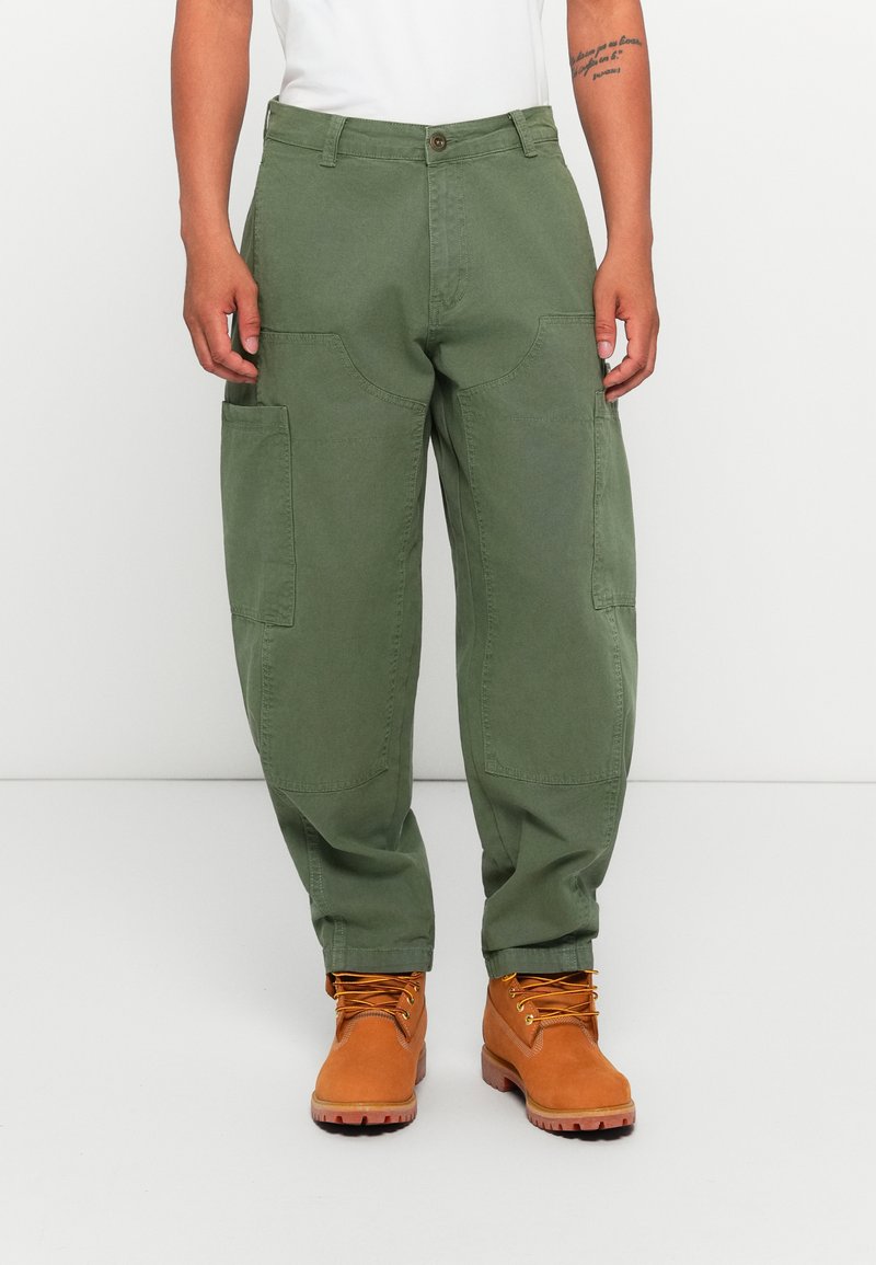 Person wearing loose olive green cargo pants and tan lace-up work boots, standing against a plain white background.