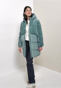 Padded green jacket z kapturem, z pikowanym wzorem, z przodu na zamek, z kieszeniami po bokach, w połączeniu z ciemnymi spodniami i trampkami.