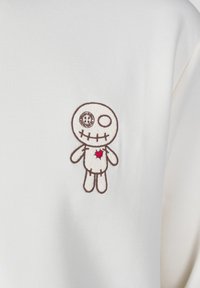 Top de tejido blanco con un diseño bordado de muñeco vudú con contornos marrones, un detalle de calavera y ojo, y un corazón rojo en el pecho.