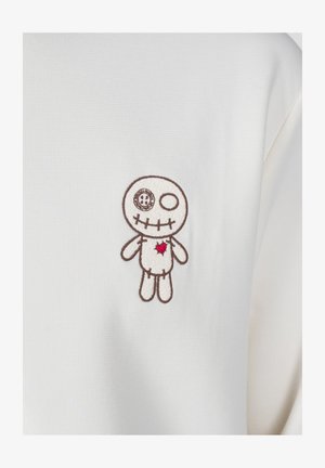 Biała bluzka z materiału z haftowanym wzorem lalki voodoo, brązowymi konturami, detalem czaszki i oka oraz czerwonym sercem na klatce piersiowej.