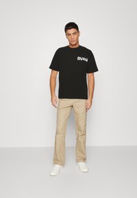 Black cotton t-shirt with front logo, paired with beige chinos and white sneakers. Model stands against a plain background.