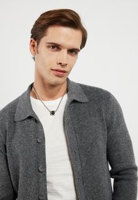 Gråstrikket cardigan med kragehals, knappesfront og ribbet tekstur, båret over en hvit T-skjorte. Sølvkjede synlig.