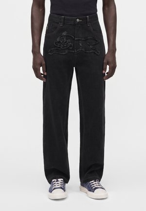 Personne portant un jean noir à jambes larges avec un motif animal texturé à l’avant et des baskets bleues avec des lacets blancs.