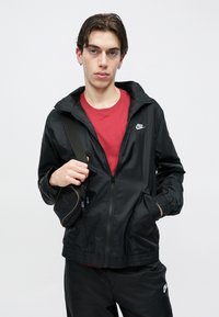 Fiatal férfi fekete Nike dzsekiben és nadrágban, piros pólóval, egyik vállán fekete oldaltáska pántját tartva.