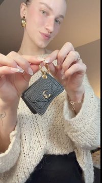 Mustast tekstureeritud nahast kaarditasku kuldsete detailide ja C-kujulise aktsendiga. Seda hoiab inimene, kes kannab kreemikat kootud kampsunit, neutraalses keskkonnas.