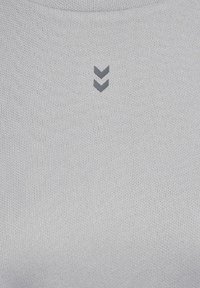 Helles graues Sportoberteil aus strukturiertem Material mit einem kleinen dunkelgrauen Chevron-Logo, das zentral auf der Brust platziert ist.