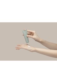 Vihreä putki puristaa valkoista voidetta kädelle. Tausta on neutraali beige, joka korostaa tuotetta ja sen rakennetta.