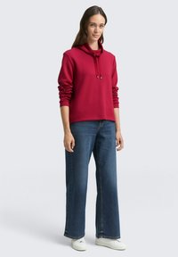 Roter Kapuzenpullover mit lässigem Schnitt, gerippten Bündchen und Saum, kombiniert mit weitgeschnittenen blauen Jeans und weißen Sneakers.