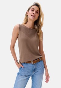 Bruine mouwloze top met een textuurpatroon op de borst, gecombineerd met lichtblauwe jeans en een bruine riem. Soepele stof, casual ontwerp.