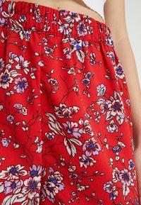 Červené květinové kalhoty s elastickým pasem, zdobené bílými a fialovými květinami na texturované tkanině. Lehký a volný střih.