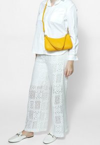 Witte kanten wijde broek met een ingewikkeld diamantpatroon, gecombineerd met een losse witte blouse met knopen en een felgele schoudertas.