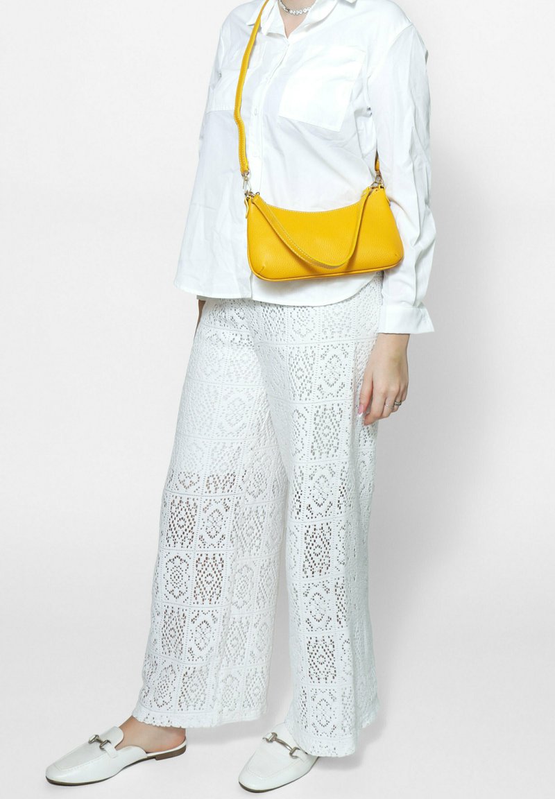 Witte kanten wijde broek met een ingewikkeld diamantpatroon, gecombineerd met een losse witte blouse met knopen en een felgele schoudertas.