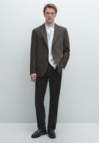 Brauner strukturierter Blazer über einem weißen Hemd, kombiniert mit dunklen Hosen und schwarzen Schuhen. Verfügt über zwei Knöpfe und eine taillierte Passform.