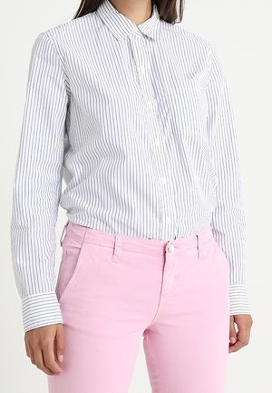 Kvinde iført en hvid skjorte med tynde lodrette sorte striber og lyserøde bukser, vist fra nakke til hofter.