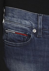 Jeans de denim azul oscuro con un bolsillo frontal que tiene un acento de franjas blanca y roja, herrajes de botón de metal y un diseño minimalista.