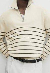 Pull côtelé crème à fermeture éclair courte avec des rayures horizontales sombres, doté d'un détail zippé au col. Associé à un pantalon clair et une ceinture noire.