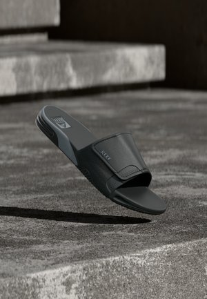 Černá slide sandála se širokým páskem a strukturovanou stélkou ležící na betonovém schodu venku.