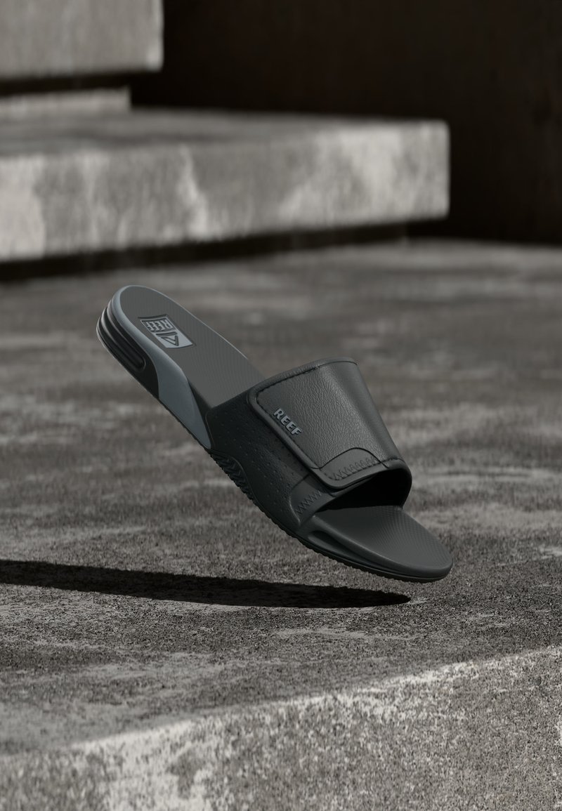 Sandale slide noire avec une large bride et une semelle texturée reposant sur une marche en béton à l'extérieur.