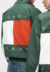 Δύο άτομα φορούν μπουφάν σε μπλοκ χρωμάτων πράσινο, άσπρο και κόκκινο με την επιγραφή "Tommy Jeans" κεντημένη στην πλάτη και στα μανίκια.