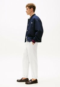 Giacca bomber blu navy con bottoni a pressione bianchi e polsini a coste, abbinata a pantaloni bianchi larghi e mocassini marroni.