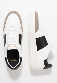 Vita lädersneakers med svarta och grå mockadetaljer, perforerat tåbox, platta snören och texturerad vit gummisula. Guld logotypdetalj.