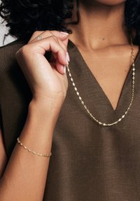 Złoty naszyjnik i bransoletka z owalnymi ogniwami. Naszyjnik opada na brązowy top z dekoltem w serek. Gładka struktura całości.