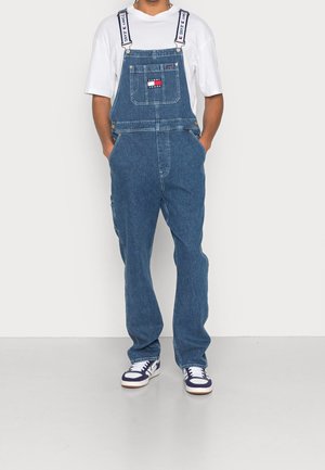 Osoba ubrana w niebieskie dżinsowe ogrodniczki Tommy Jeans z białymi, regulowanymi paskami z logo, białą koszulkę i białe oraz granatowe trampki, stojąca z rękami w kieszeniach.