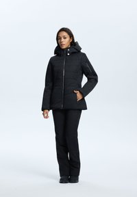 Schwarze isolierte Jacke mit Reißverschluss, abnehmbarer Kapuze und Seitentaschen. Getragen mit schwarzen Hosen und Stiefeln, die eine glatte Textur aufweisen.