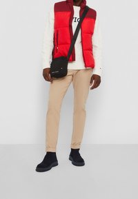 Hombre con chaleco acolchado rojo, suéter blanco de punto cable, pantalones beige, botas Chelsea negras, llevando un bolso cruzado negro con logo.