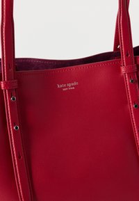 Κόκκινη δερμάτινη τσάντα ώμου με ρυθμιζόμενους ιμάντες, με λογότυπο "kate spade NEW YORK" χαραγμένο σε ασημί στο μπροστινό μέρος.