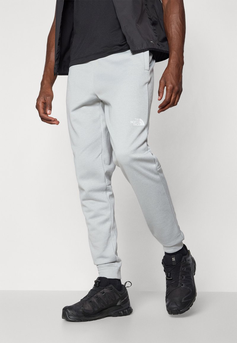 Un pantalon de survêtement gris clair avec des jambes fuselées, des poignets élastiques et un logo blanc sur la cuisse, associé à des chaussures de sport noires avec une tige texturée.