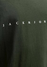 Olivenfarbene T-Shirt mit dem Text "JACK&JON" in Weiß, der sich in der oberen linken Ecke befindet. Der Stoff hat eine glatte Textur.