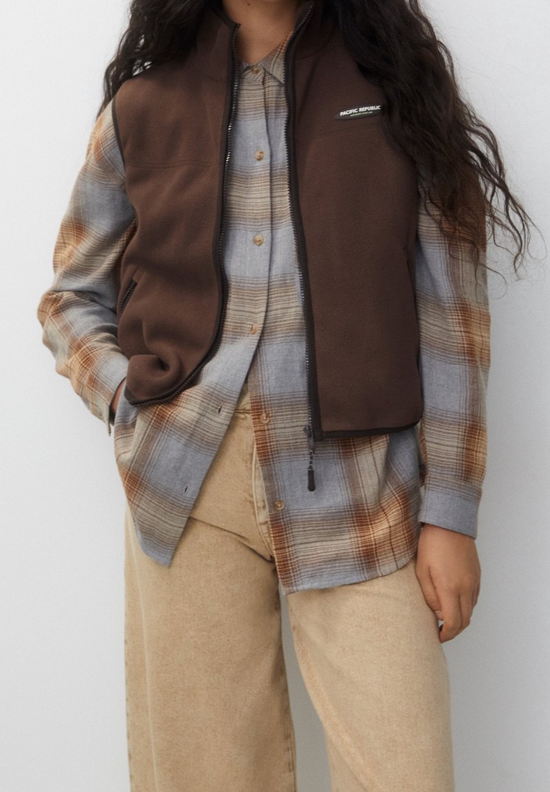 Personne portant un pantalon beige, un gilet sans manches en polaire marron et une chemise à carreaux gris et orange à manches longues avec boutons.