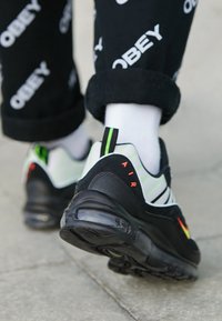 Černobílé tenisky s greenskými akcenty, které mají texturovanou černou podrážku, pruhy po stranách a červený detail "AIR" na patě.