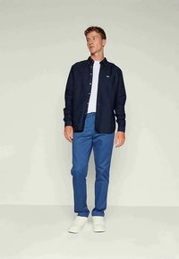 Cămașă cu mâneci lungi de culoare bleumarin, cu nasturi, purtată peste un tricou alb, asortată cu pantaloni albaștri și adidași albi; prezintă logo-ul Lacoste pe cămașă.