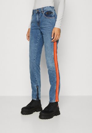 Λεπτά μπλε τζιν με πορτοκαλί και λευκές ρίγες στο πλάι, φερμουάρ στον αστράγαλο, φορεμένα με μαύρες χοντρές μπότες και λευκό μακρυμάνικο μπλουζάκι.