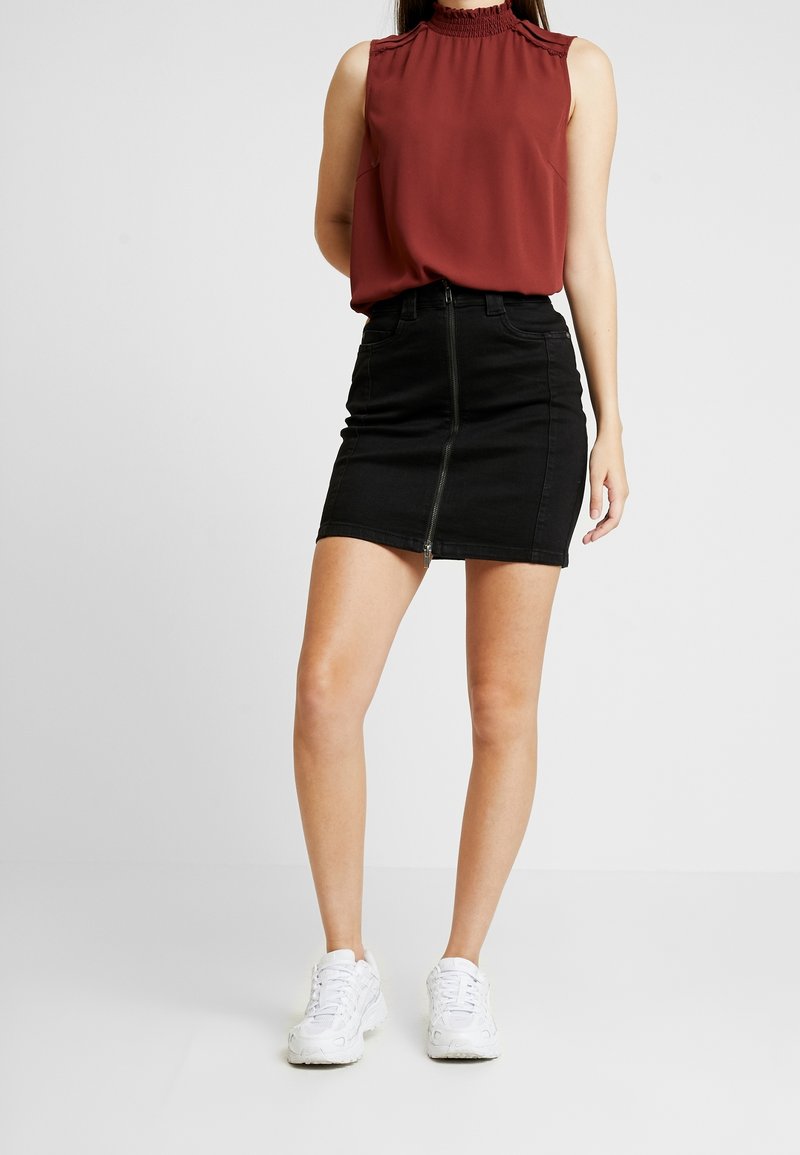 Femme portant un chemisier rouge sans manches, une mini-jupe noire avec fermeture éclair devant et des baskets blanches, debout devant un fond uni.