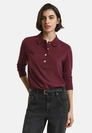 Woman wearing a long-sleeve burgundy polo shirt with gold buttons, tucked into high-waisted black jeans with a black belt.